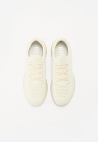 Light beige athletic shoes with a textured mesh upper, flat laces, and a cushioned sole. Features a logo on the insole and a minimalist design.