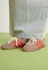 Person som bär ljusblå jeans och bruna mockaskor med röda ränder och crème-färgade snören, står på grön matta.