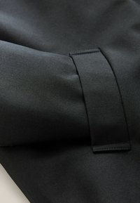 Must have suju tekstuuriga must kangas, millel on volditud osa ja nähtav õmmeldud rihma detail.