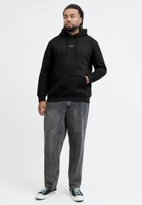 Mężczyzna ubrany w czarną bluzę z kapturem, szare jeansy typu washed oraz czarne trampki, stojący z jedną ręką w kieszeni na jednolitym białym tle.