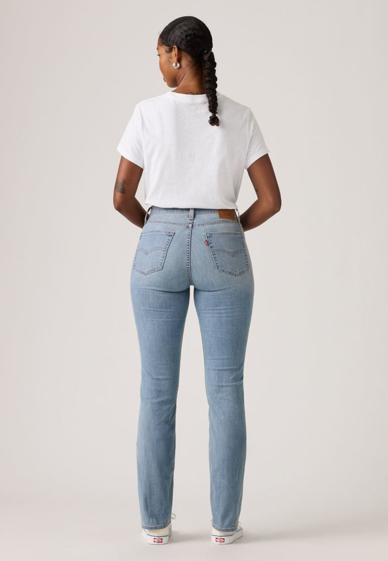 Femme aux cheveux tressés portant un t-shirt blanc, un jean taille haute bleu clair et des baskets blanches, debout de dos les mains sur les hanches.