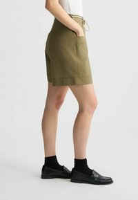 Shorts verde oliva con vita regolabile, tasche laterali e orlo piegato. Abbinati a scarpe nere e calzini scuri. Texture del tessuto liscia.