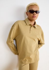 Polo a manica lunga color khaki con colletto, con un discreto logo Nike. Il modello indossa occhiali da sole eleganti, abbinati a pantaloni khaki.