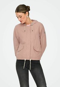 Femme portant une veste à capuche rose poussiéreux avec des poches frontales et des cordons de serrage, associée à un jean gris foncé, regardant vers le bas les cheveux attachés en arrière.