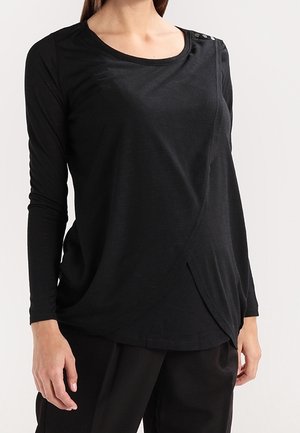 Žena má na sobě černý top s dlouhým rukávem ve stylu zavinovacího střihu s knoflíky u výstřihu a volné černé kalhoty.