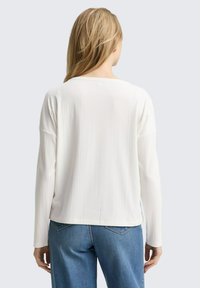 Vrouw gezien van achteren, in een wit, lange mouw ribbelshirt en blauwe jeans, tegen een effen lichte achtergrond.