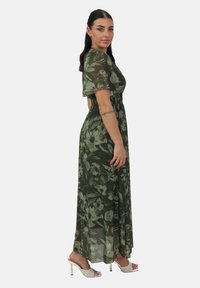 Grünes Blumen-Maxikleid mit kurzen Ärmeln, elastischem Bund und transparenter Stoffqualität. Es hat ein fließendes Design und wird mit weißen Absatzsandalen kombiniert.