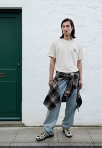 Maglietta beige chiaro con grafica, jeans larghi azzurri chiari, camicia a quadri bianca e nera legata in vita e sneakers con motivo a fantasia.