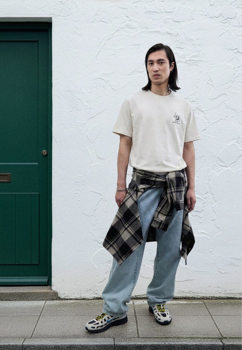 Maglietta beige chiaro con grafica, jeans larghi azzurri chiari, camicia a quadri bianca e nera legata in vita e sneakers con motivo a fantasia.