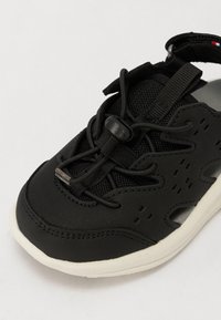 Μαύρο αθλητικό παπούτσι από πλέγμα και καουτσούκ, με υφή στην επιφάνεια, ελαστικούς κορδόνες και λευκή σόλα. Περιλαμβάνει ρυθμιζόμενο λουρί.