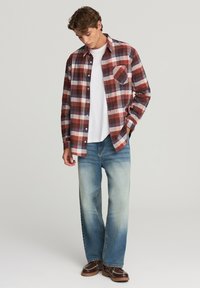 Camicia flanella a quadri rossa, blu e crema con maniche lunghe e tasca frontale, abbinata a jeans blu scoloriti e sneakers marroni.