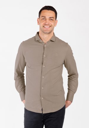Camisa de manga larga en un verde oliva claro, diseño con botones en el frente, textura suave, logo sutil en el pecho, combinada con jeans oscuros.