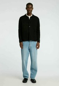 Fekete gombos ing, hosszú ujjal, fehér póló felett viselve, világoskék farmernadrággal és fekete lapos talpú cipővel párosítva.