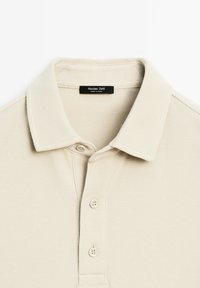 Beige polo tričko s texturovanou tkaninou. Má skládací límeček, zapínání na dvě knoflíky a malou černou značkovou etiketu na výstřihu.