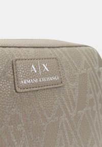 Borsa beige strutturata con motivo in rilievo. Presenta un'etichetta con logo A|X, con il nome del marchio "Armani Exchange" in lettere bianche.