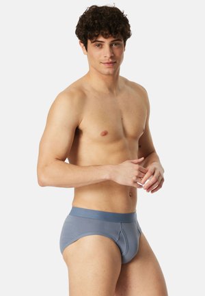 Junger Mann mit lockigem Haar, der graue Unterhosen trägt, vor einem schlichten hellen Hintergrund steht und mit einem leichten Lächeln in die Kamera schaut.