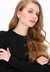 Zwarte gebreide cardigan met een gestructureerd zigzagpatroon, voorzien van ronde bruine knopen en ribgebreide mouwen. Het model heeft lang golvend haar.