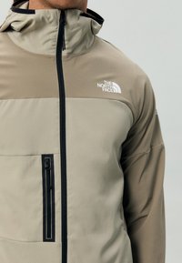 Homme portant une veste beige et marron clair avec une fermeture éclair noire et une poche poitrine, avec le logo The North Face sur la poitrine.