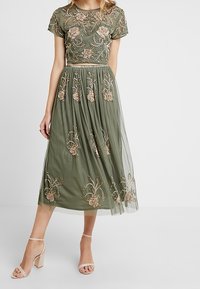 Olijfgroene midi-jurk met een beaded bloemenpatroon, doorschijnende overlay, korte mouwen en een getailleerde bovenkant. Gecombineerd met naaktkleurige sandalen met bandjes.