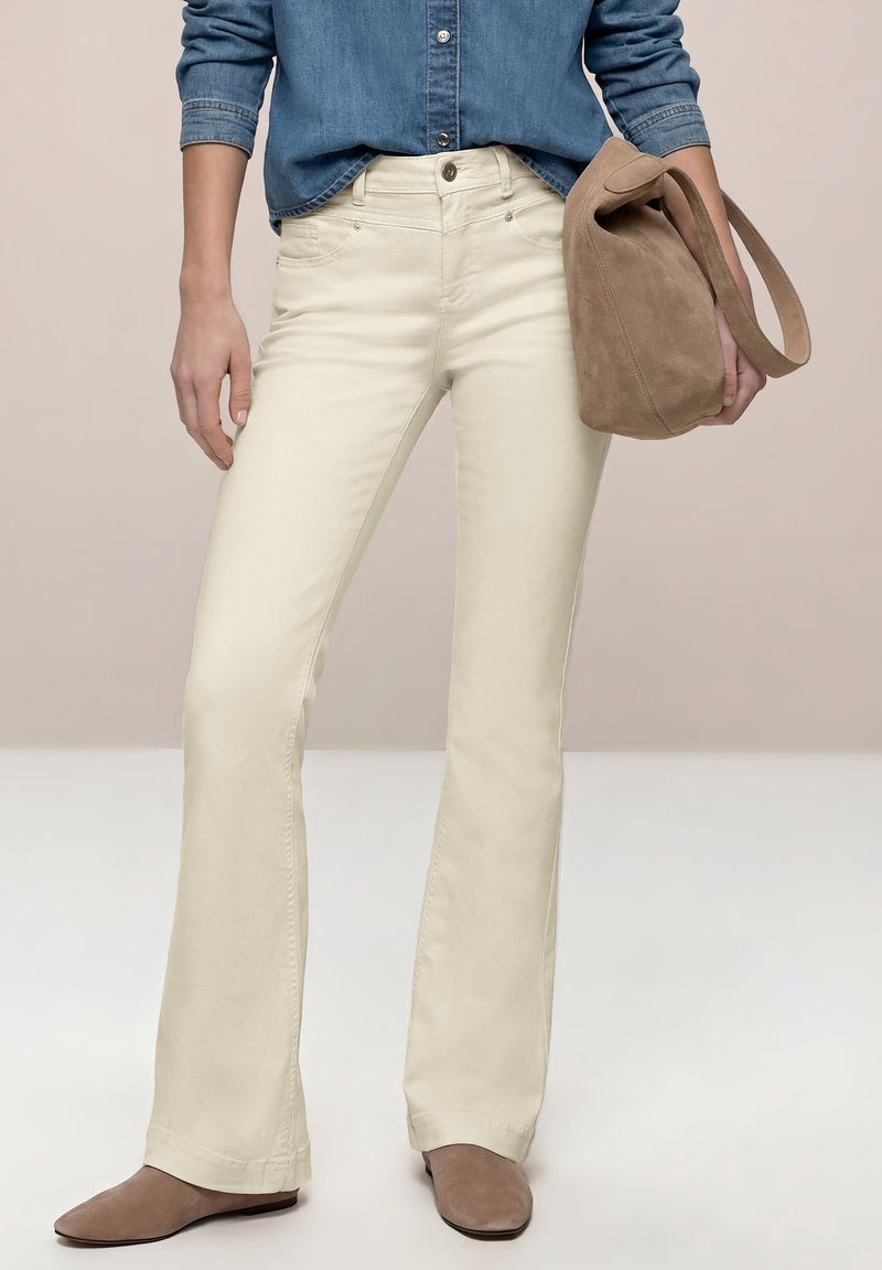 Persona con pantalones acampanados color crema, zapatos de ante marrón, camisa de mezclilla, sosteniendo un bolso de ante topo en una mano.