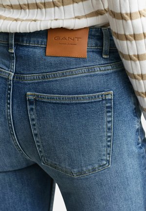 Denim jeans back pocket with brown GANT brand label, worn with a white and beige striped ribbed sweater.