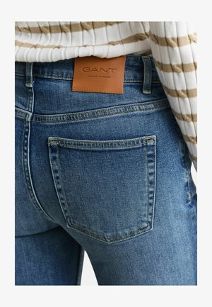 Poche arrière d'un jean en denim avec une étiquette de marque GANT marron, portée avec un pull à côtes rayé blanc et beige.
