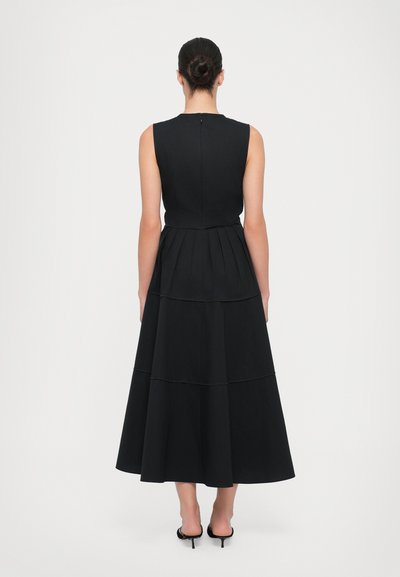 Vestido negro sin mangas con un corpiño ajustado y una falda acampanada y fluida en capas. Presenta detalles de costura sutiles y un cierre de cremallera en la parte posterior.