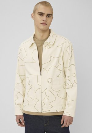 Jeune homme aux cheveux blonds courts portant une veste à fermeture éclair beige à motifs floraux sur une chemise marron et un pantalon foncé, sur un fond gris uni.