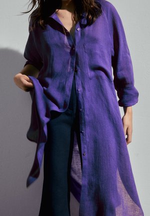 Femme portant une chemise longue semi-transparente violette par-dessus un pantalon foncé, tenant une partie de la chemise d'une main devant un fond uni.