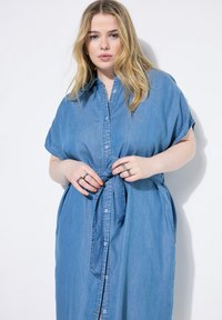 Denim mekko lyhyillä hihoilla, napit edessä, solmittava vyötäröyksityiskohta ja vaalean sininen väri. Sileä pinta, jossa on hienovaraisia ommelkoristeita.