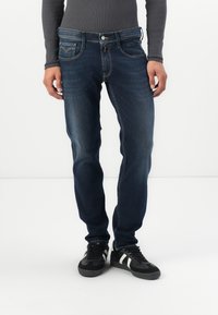 Jeans en denim bleu foncé au design ajusté, cinq poches, décoloration subtile et coutures contrastées, associés à des baskets noires.