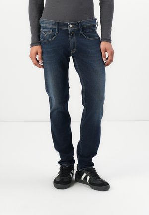 Mörkblå denimjeans med en åtsittande design, fem fickor, subtil blekning och kontrasterande sömmar, tillsammans med svarta sneakers.