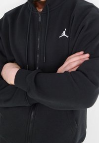 Person wearing a black zip-up hoodie with a white Jordan logo on the chest, arms crossed across the torso.