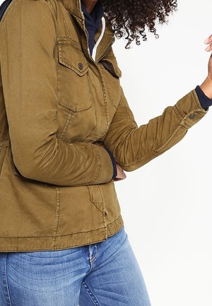 Veste zippée brun-vert avec deux poches poitrine, détails en bouton et tissu texturé. Portée sur un hoodie bleu, associée à un jean bleu.