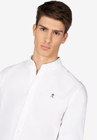 Camisa blanca de botones confeccionada en algodón, con un cuello clásico y un pequeño logo azul oscuro en el lado izquierdo del pecho. Textura suave, ajuste entallado.