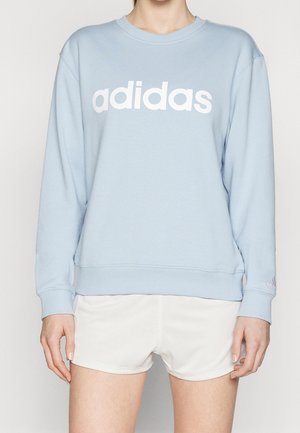 Persoon draagt een lichtblauwe Adidas trui met wit logo over de borst en witte sportshort, staand tegen een effen achtergrond.