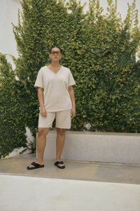 Krémovo béžový top s krátkym rukávom a výstrihom do V a zodpovedajúce šortky z mäkkej látky, doplnené o čierne sandále na klíne a žlté slnečné okuliare. Pozadie zo zelenej vegetácie.