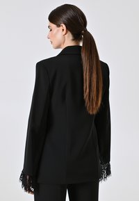 Blazer nero a maniche lunghe, dettagli in pizzo sui polsini, design strutturato, texture liscia e vestibilità su misura, visto dal retro.