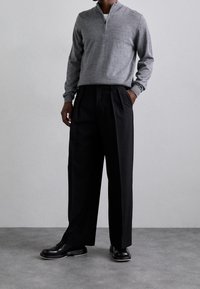 Pull gris à fermeture éclair avec un col rond, associé à un pantalon large noir et des chaussures noires sur un fond gris texturé.