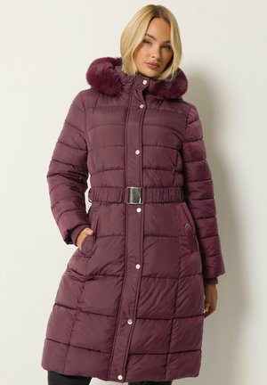 Femme portant un long manteau matelassé violet avec une capuche bordée de fourrure et une taille ceinturée, debout devant un fond clair uni.