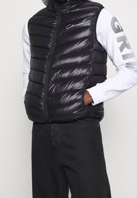 Schwarze Steppweste mit horizontalen Nähten und durchgehendem Frontreißverschluss. Das Material ist glänzend, leicht, mit hohem Kragen und einem Markenlogo.