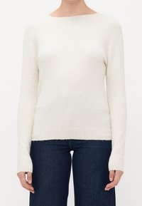 Femme portant un pull en tricot à manches longues de couleur beige clair associé à un jean bleu foncé, debout devant un fond blanc uni.