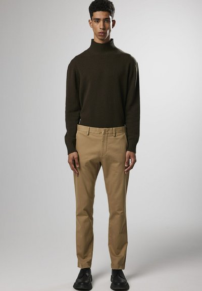 Donkergroene coltrui, beige slim-fit chinos en zwarte schoenen. Model staat rechtop en toont een eenvoudige, hedendaagse outfit.