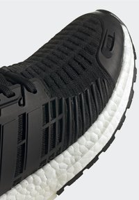 Chaussure de course en maille noire avec cage en plastique noir, semelle en mousse blanche texturée et semelle extérieure en caoutchouc noir, montrée en gros plan.