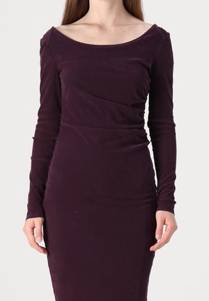 Vrouw draagt een nauwsluitende, lange mouwen, donkerpaarse fluwelen jurk met een brede, afgeronde halslijn tegen een effen achtergrond.