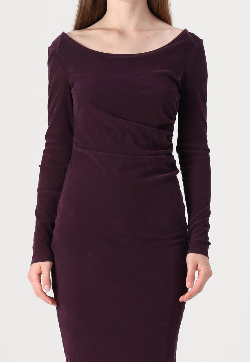 Femme portant une robe ajustée en velours violet foncé à manches longues, avec un large décolleté arrondi, sur un fond uni.