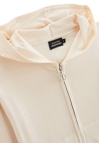Haut de survêtement beige zippé avec une texture douce et côtelée, doté d'une capuche à cordon de serrage et d'un petit zip en métal. L'étiquette indique "MONOPRIX".