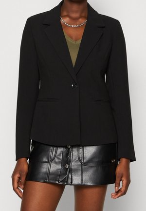 Kobieta ubrana w czarną marynarkę na jeden guzik, nałożoną na oliwkowozielony top oraz czarną skórzaną mini spódnicę z przodu zapinaną na guziki.