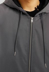 Grå huva jacka med en slät textur, med en framdragkedja och snoddar. Tyget verkar lätt och vattenavvisande.