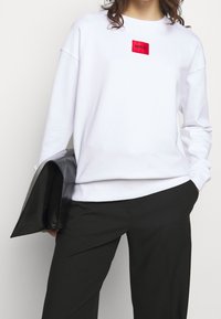 Sweat-shirt blanc avec un patch logo rectangulaire rouge. Coupe ample avec épaules tombantes, associé à un pantalon noir fuselé. Pochette noire tenue en main.